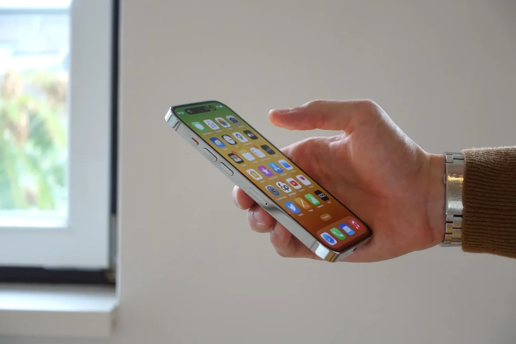 A person holding a silver iPhone with the home screen visible, demonstrating how to navigate iOS settings. The phone features the Dynamic Island and a modern interface, set against a clean, minimalist background.