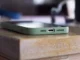 A close-up shot of an iPhone in a green protective case resting on a wooden table, highlighting the bottom charging port and speakers.