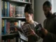 A man and woman standing in front of a bookshelf filled with manga and graphic novels, each reading a different book to compare styles.