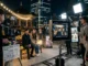 A behind-the-scenes photograph of a television production crew filming two actors on a park bench at night in Seoul. In the foreground, a large production monitor and tablets display the original webtoon panels from which the scene is adapted, with the production clapperboard confirming the source.