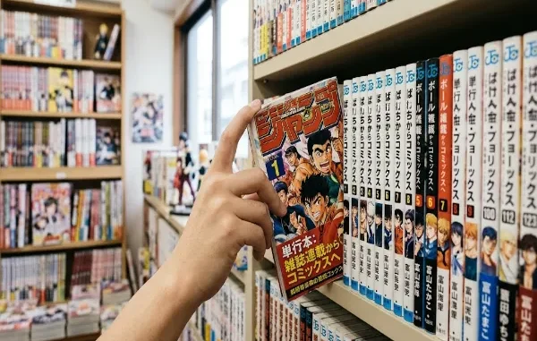 A customer's hand pulls a manga tankobon volume with colorful anime cover art from a bookshelf in a bookstore filled with organized manga volumes.